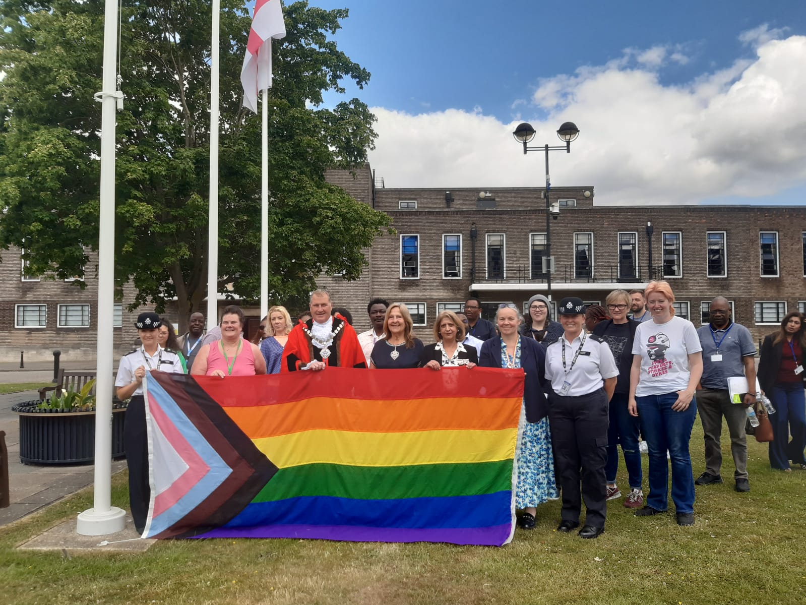 A diverse group of people stands outdoors holding a Progress Pride flag, posing and smiling in front of a flagpole and a building on a partly cloudy day.