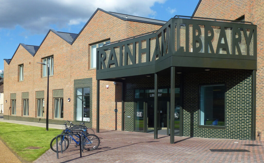 Modern brick building with large windows and a metal sign reading Rainham Library above the entrance. There are bicycle racks with bikes in front and a paved path leading to the doorway.