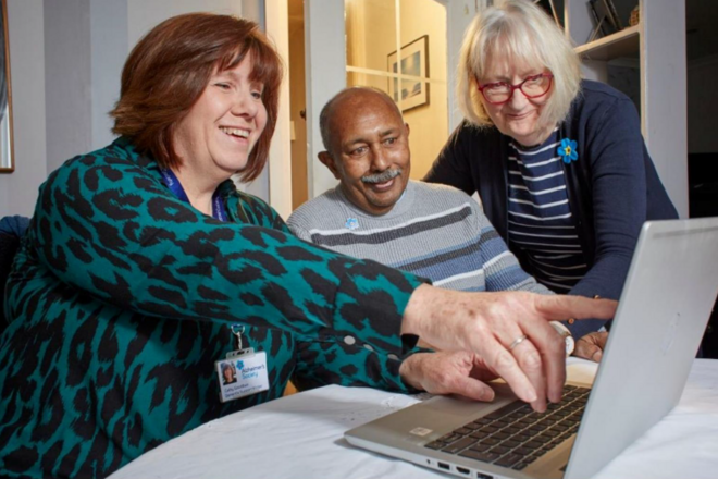 Three smiling adults sit together at a table, looking at a laptop. One woman points at the screen while the man and the other woman watch, appearing engaged and cheerful.