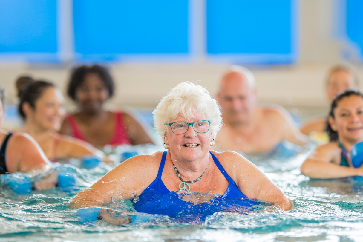 A group of people are engaged in a water exercise class inside an indoor swimming pool. They are using foam dumbbells to perform aquatic workouts.