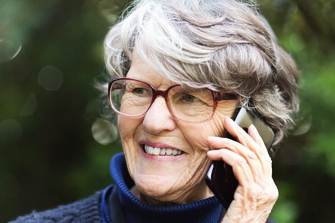 A lady with grey hair and brown eye glasses smiles while holding a smartphone to her ear.