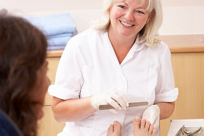 A smiling lady wearing white scrubs uses a nail file on a clients toes.