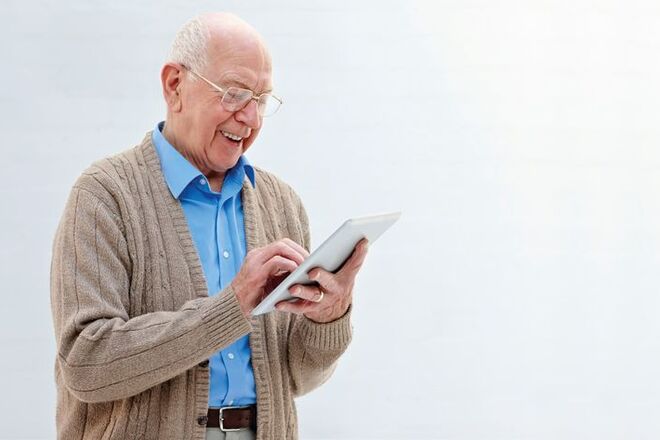 An older man stands holding a tablet. He is looking down at the tablet typing while smiling.