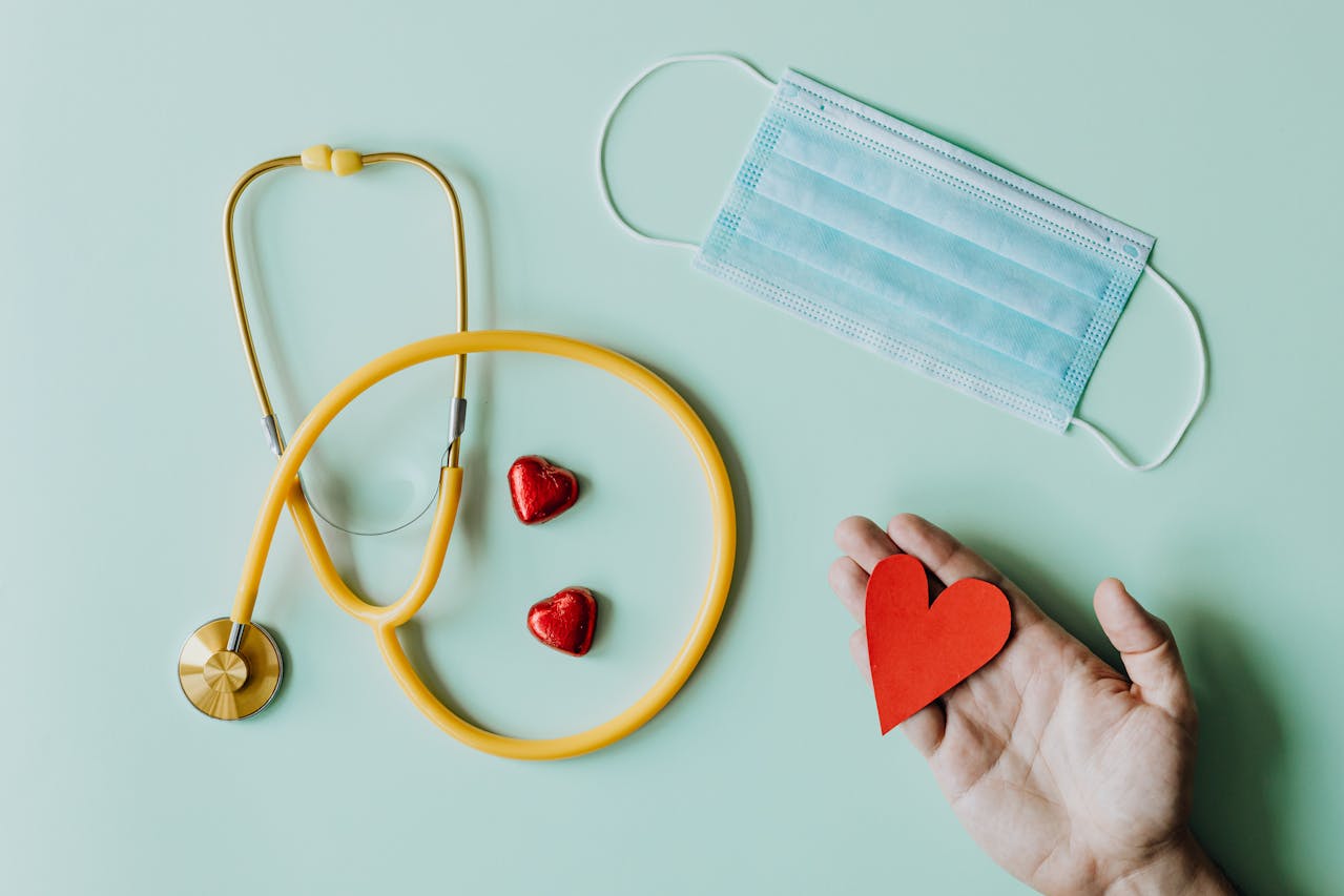Flat lay of yellow stethoscope, red hearts, blue mask, and hand holding a red paper heart on light green background.