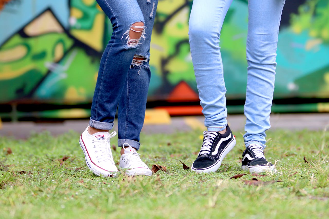 Two people standing on grass in front of a graffiti wall. One wears ripped blue jeans and white Converse; the other wears light blue jeans and black Vans