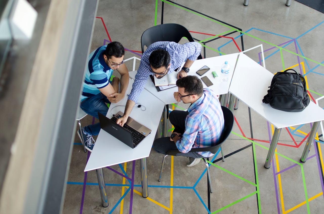 Overhead view of four people discussing around white tables with a laptop, notebooks, and a backpack; colorful tape lines decorate the floor.