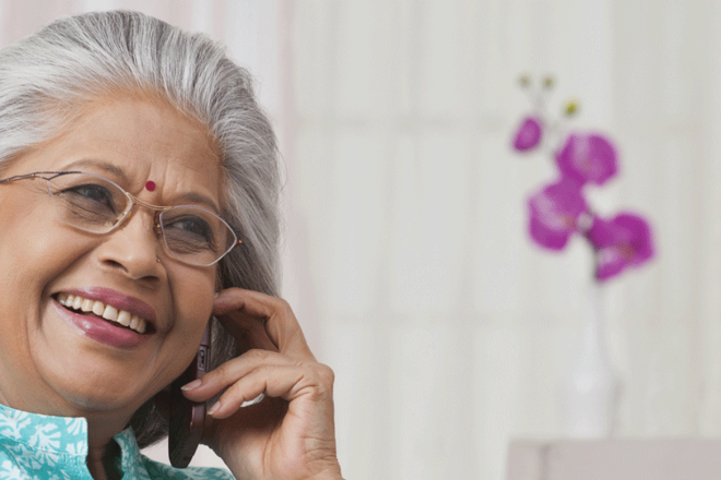 Elderly person with gray hair holding a phone with purple flowers in a vase on a table behind.