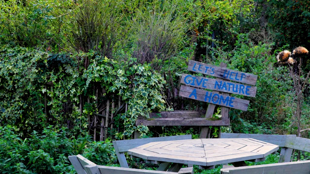A garden filled with green leaves with a wooden table and chairs in the middle. There is a sign at the back saying 'let's give nature a home'