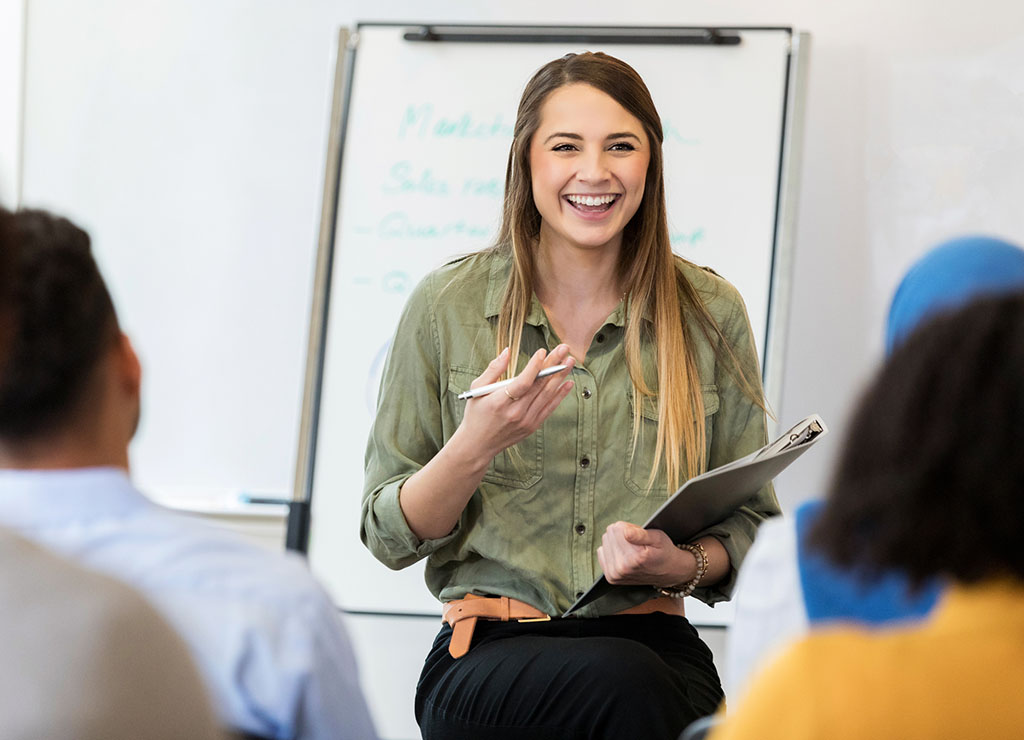 Lady smiling holding a clipboard. She is in front of a large whiteboard.
