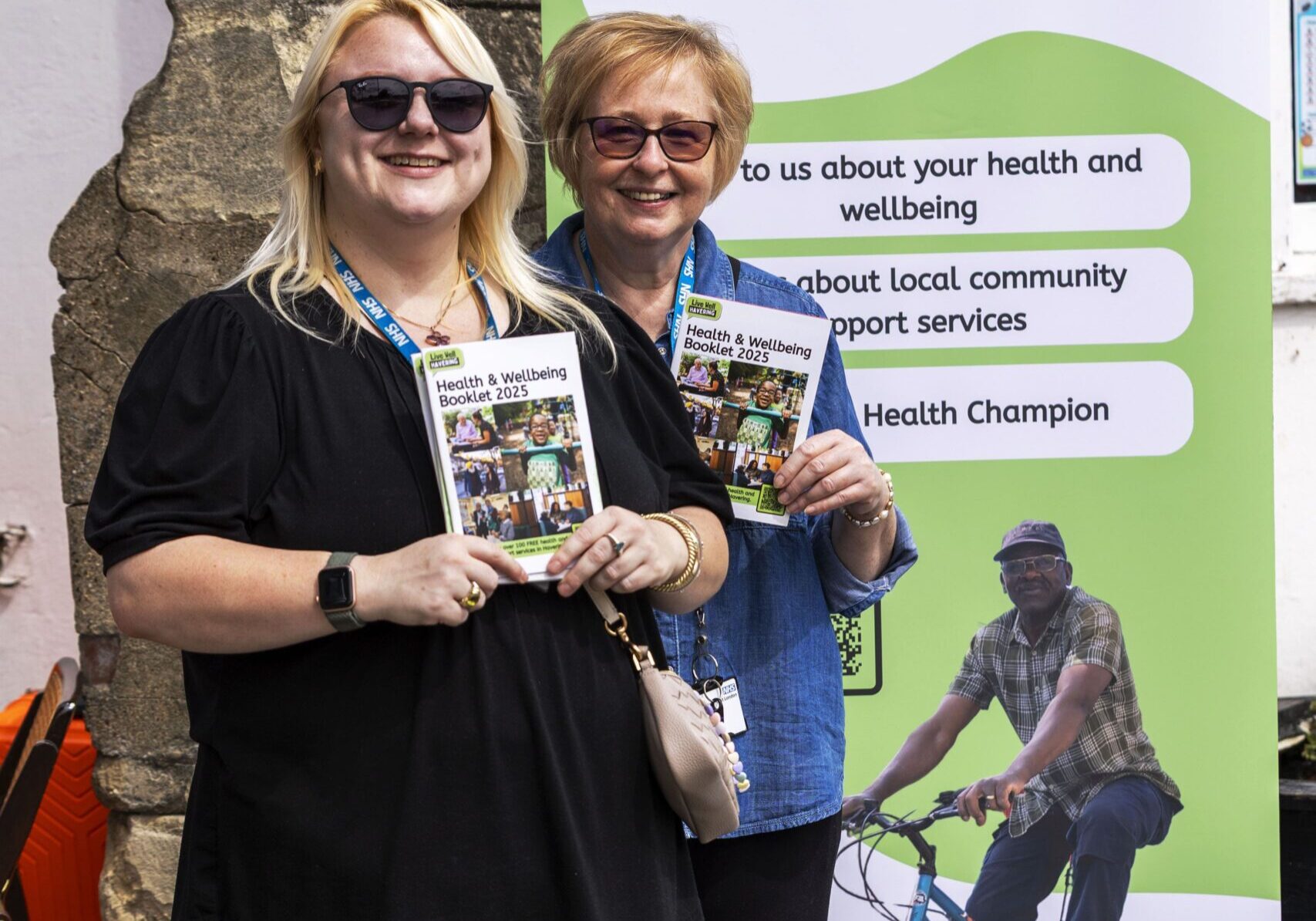 Two women stand smiling in front of a Live Well Havering banner, each holding informational leaflets. The banner promotes health, wellbeing, and community support services.