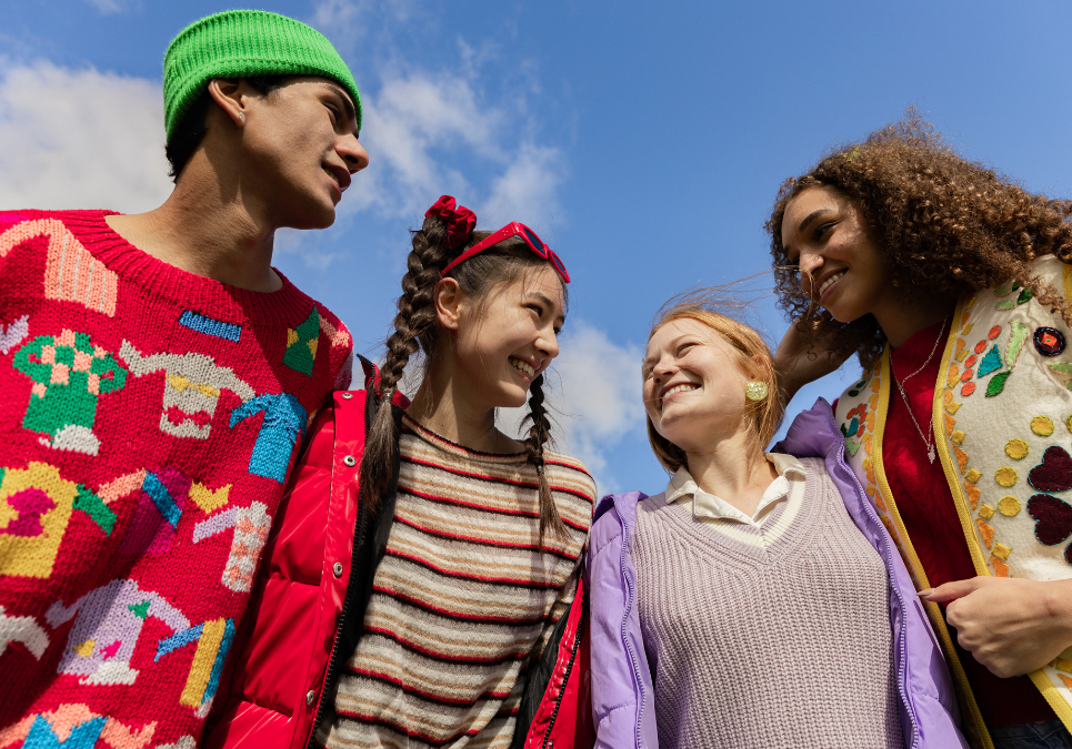 Four teenagers smiling in colour clothing stand outdoors under a blue sky.
